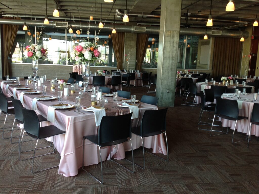 A spacious banquet hall with tables covered in pink tablecloths, black chairs, elegant floral centerpieces, candle decorations, and large windows letting in natural light. Hanging lights create a warm ambiance.