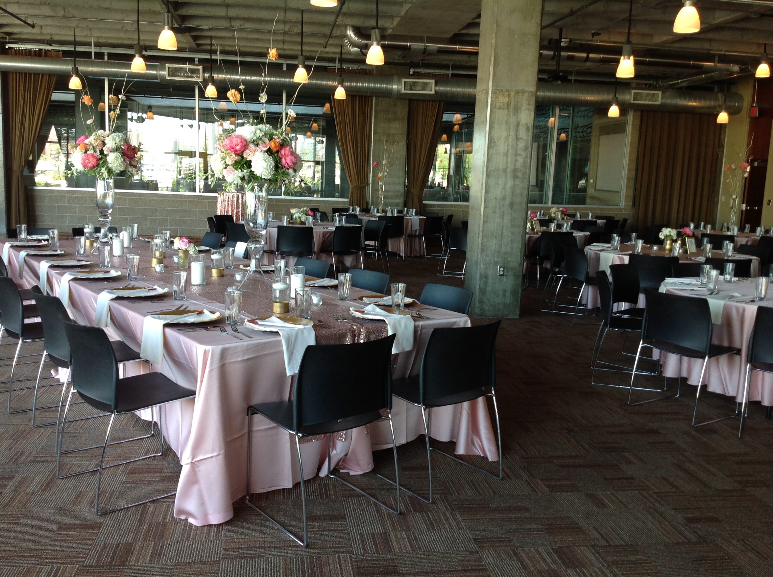 A spacious banquet hall with tables covered in pink tablecloths, black chairs, elegant floral centerpieces, candle decorations, and large windows letting in natural light. Hanging lights create a warm ambiance.