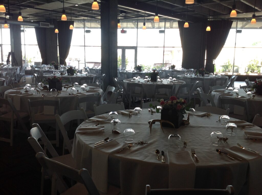 A large, elegant banquet hall set up for an event with round tables covered in white tablecloths, neatly arranged chairs, glassware, cutlery, and floral centerpieces. Hanging lights illuminate the spacious room.