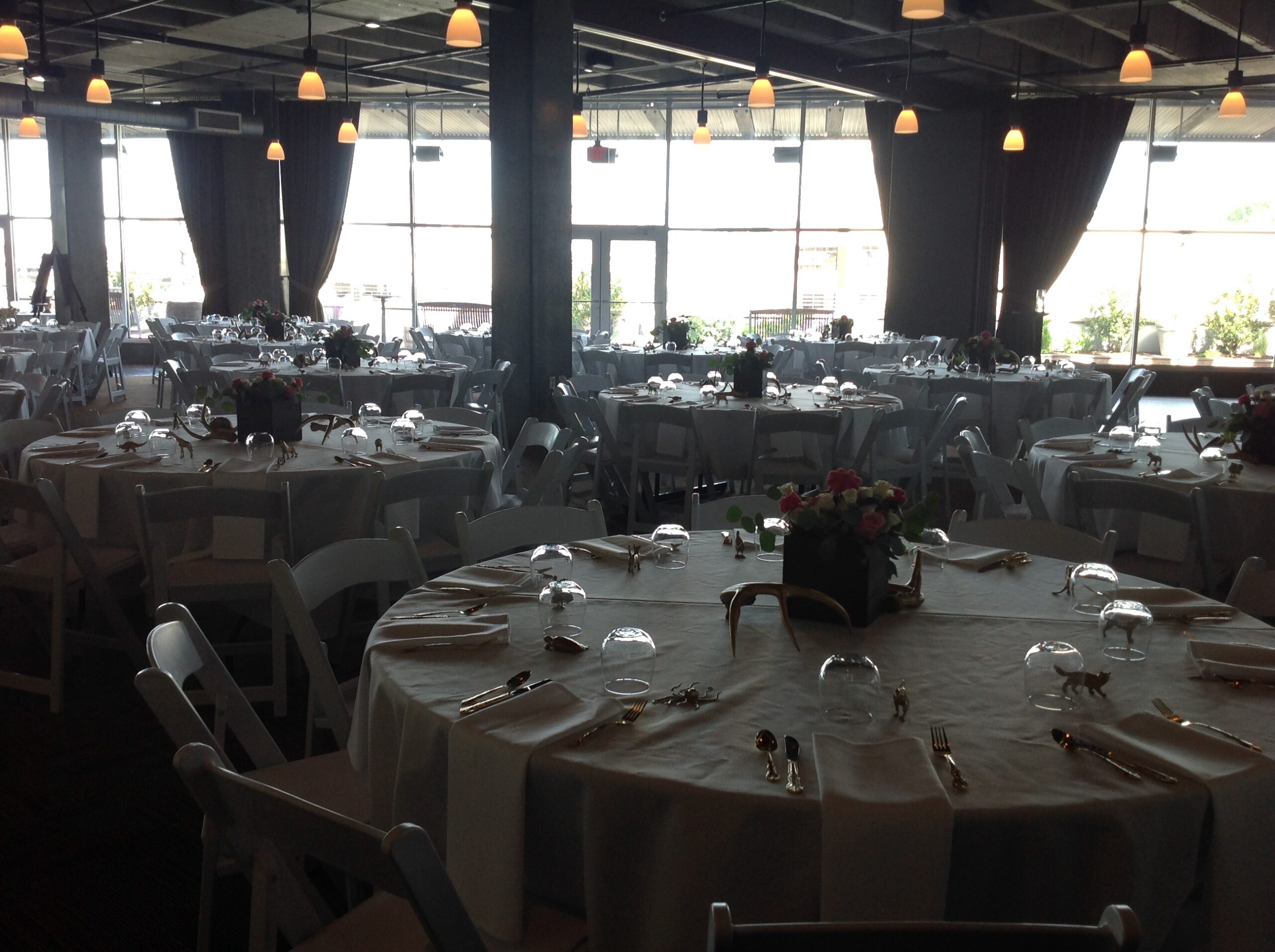 A large, elegant banquet hall set up for an event with round tables covered in white tablecloths, neatly arranged chairs, glassware, cutlery, and floral centerpieces. Hanging lights illuminate the spacious room.