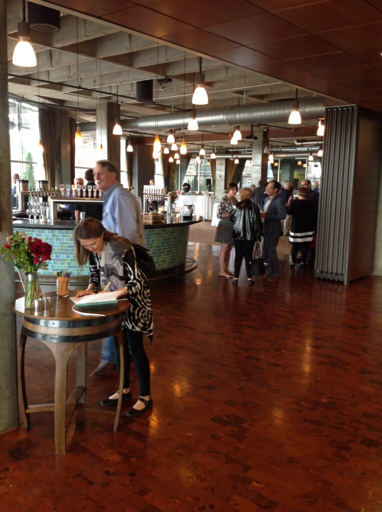 A modern, spacious cafe with a wooden floor and curved bar area. Several people stand and chat in groups, while one person writes at a small round table with flowers and pencils near the front. Warm ceiling lights illuminate the scene.