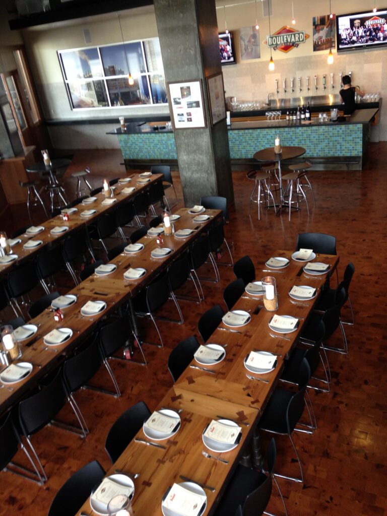 Modern restaurant interior with long wooden tables set with plates, menus, and glasses, black chairs arranged neatly; bar area with beer taps and TVs visible in the background.
