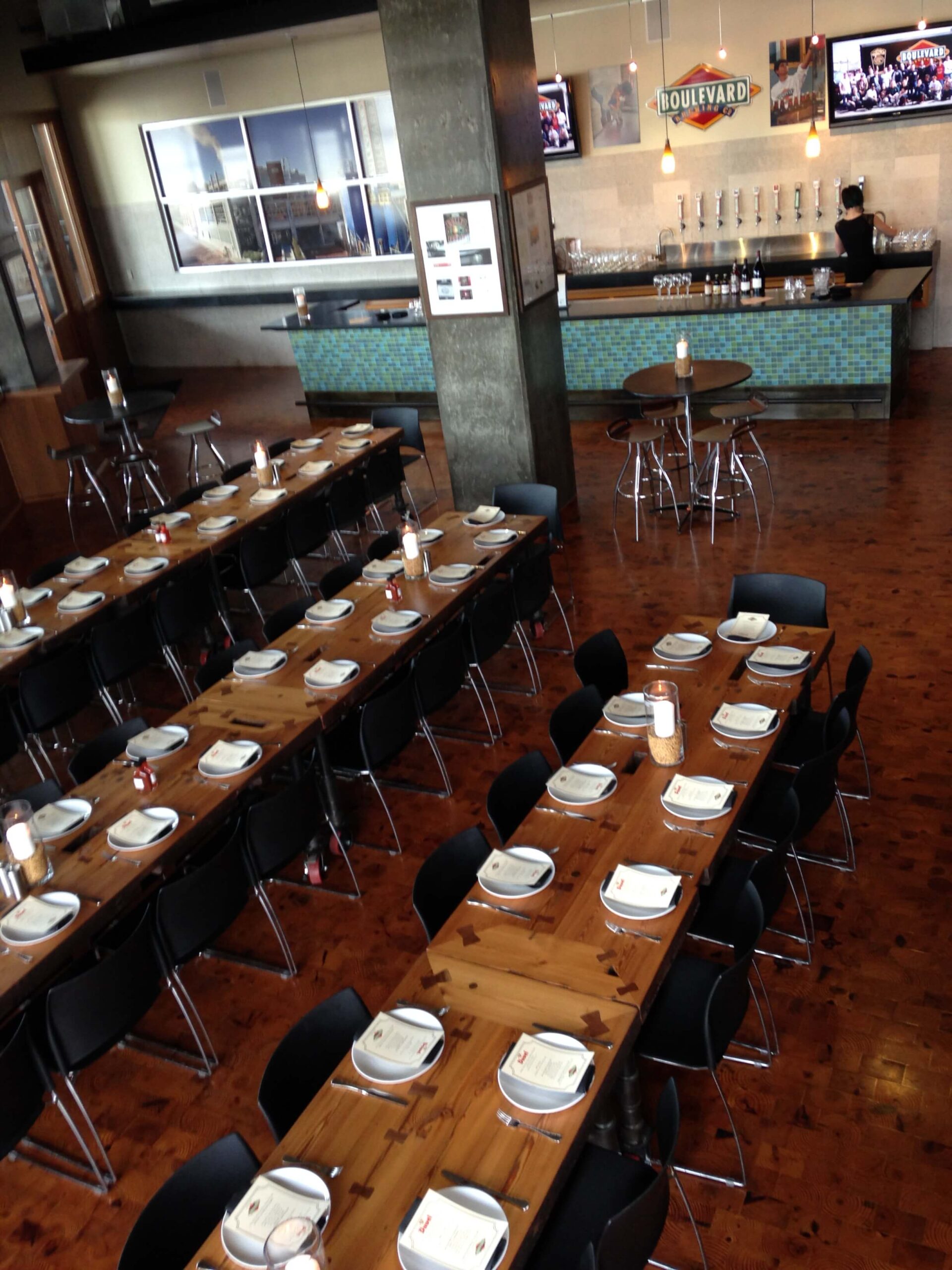 Modern restaurant interior with long wooden tables set with plates, menus, and glasses, black chairs arranged neatly; bar area with beer taps and TVs visible in the background.