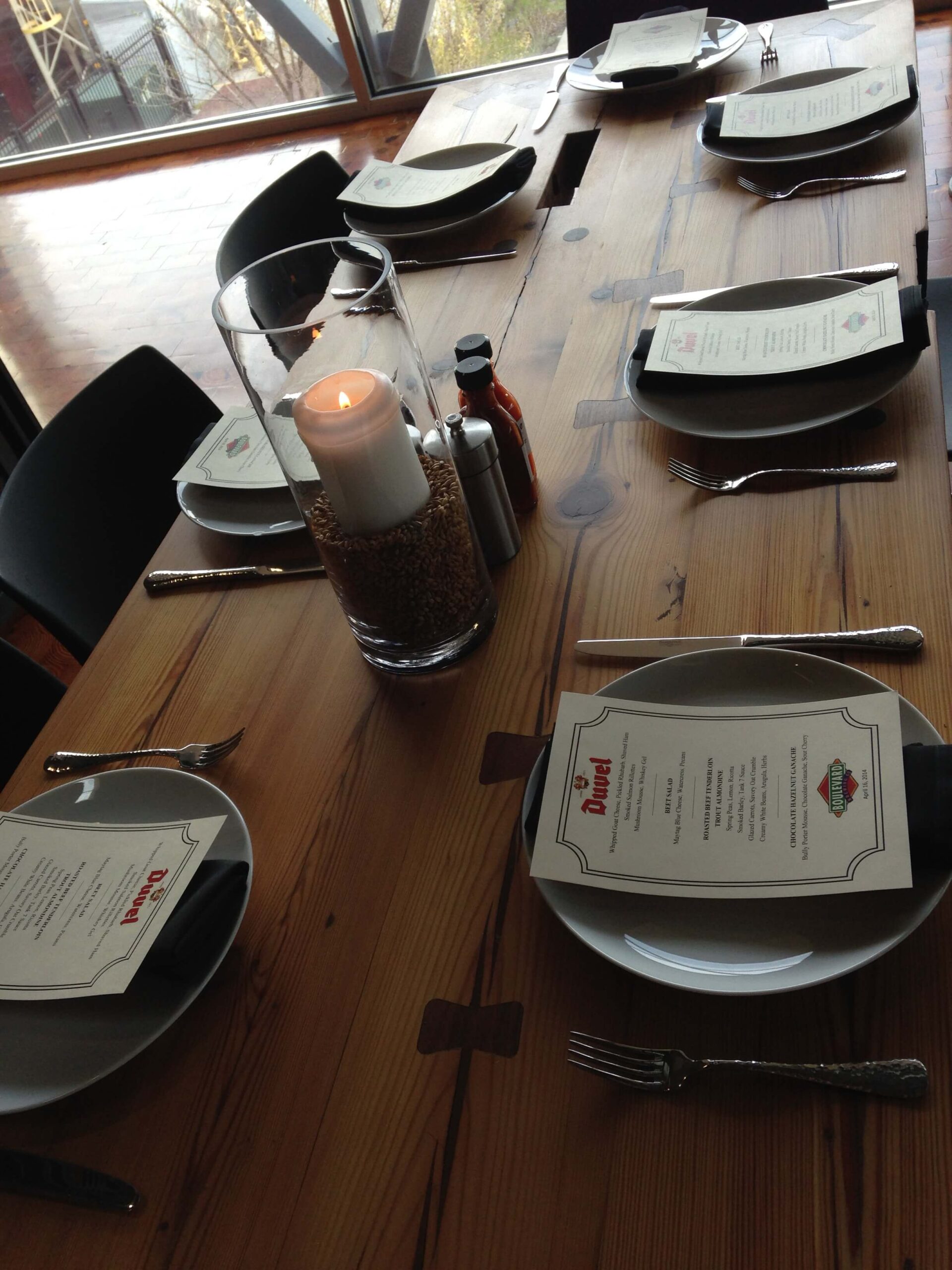 A wooden dining table set for six with plates, forks, knives, menus, a glass candle centerpiece, salt and pepper shakers, and a small bottle of hot sauce. The table is near a large window.