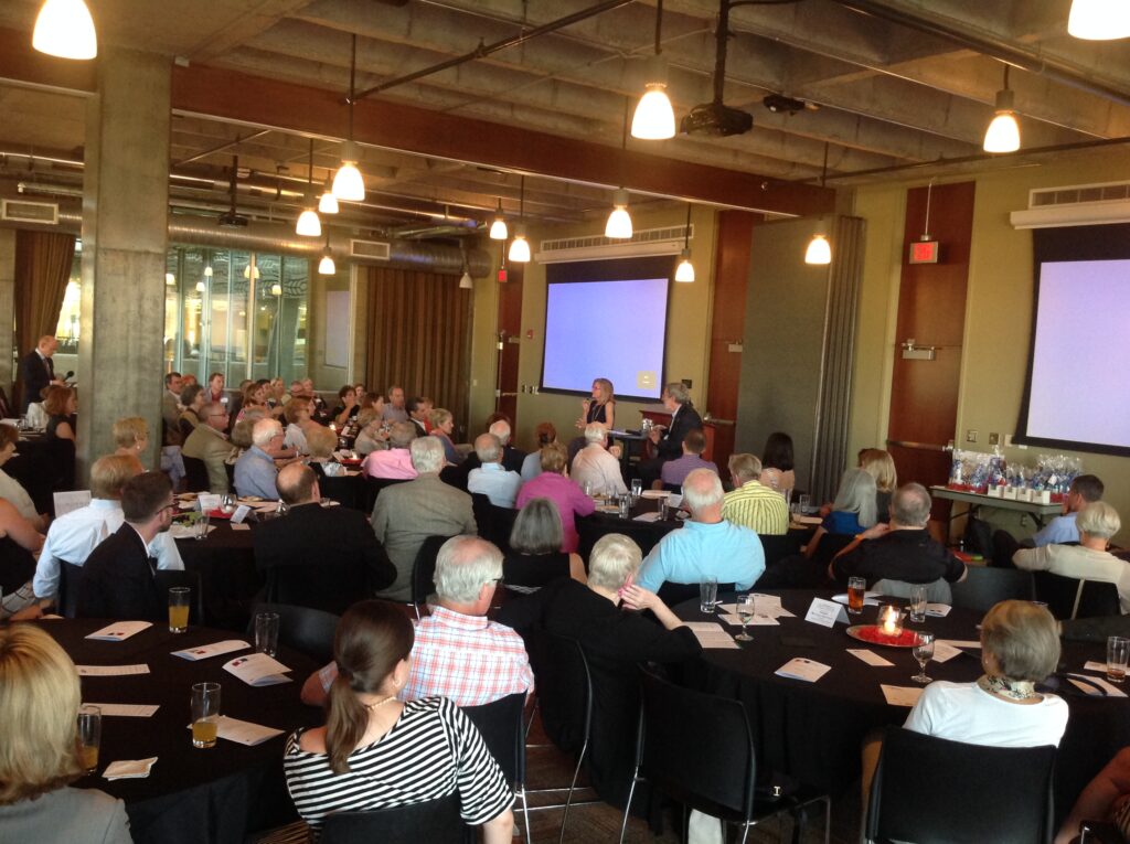 A large group of people sit at round tables in a modern event space, attentively watching a presentation at the front, where two speakers stand near screens displaying blue slides.