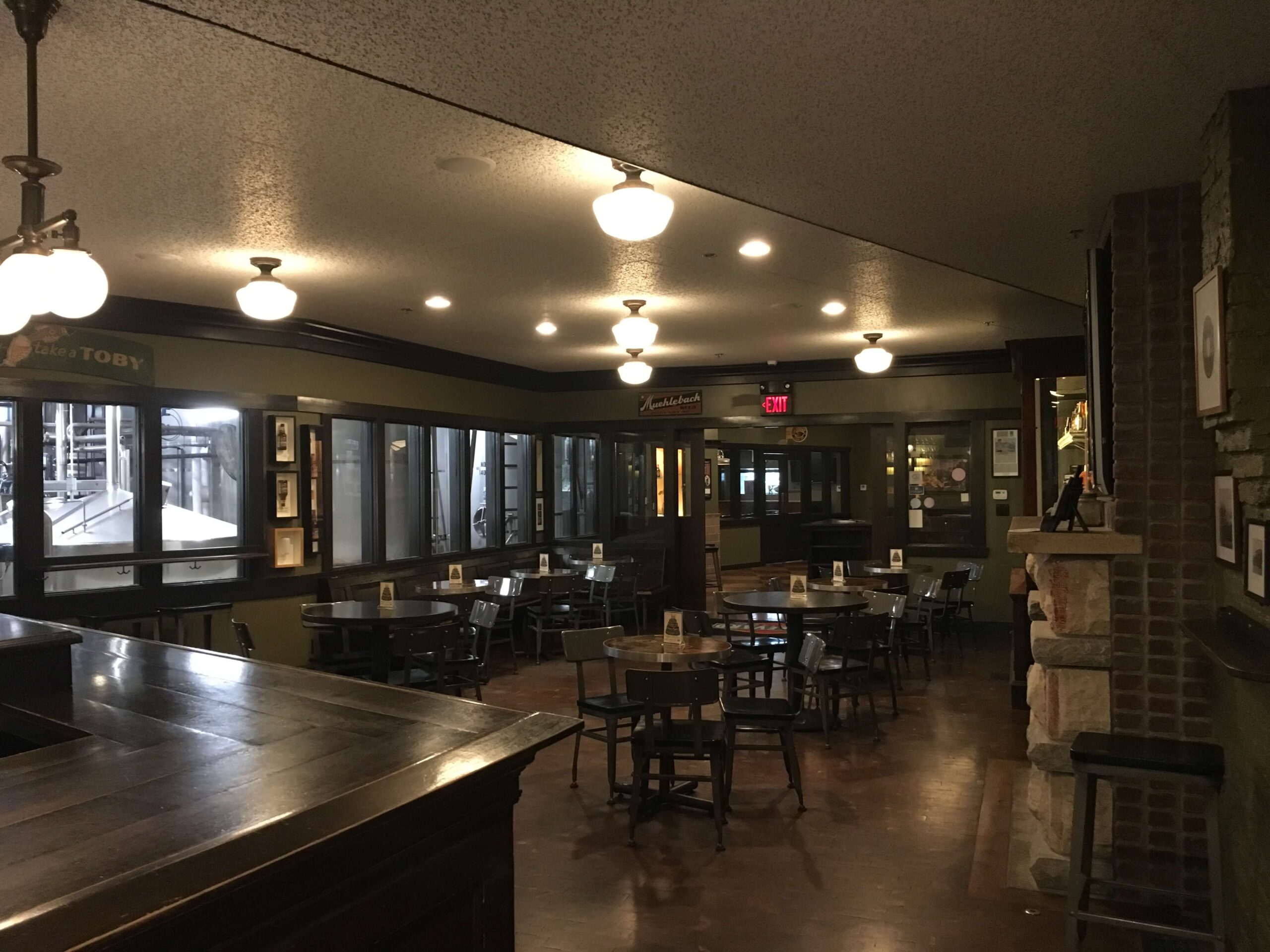 A dimly lit, empty restaurant or pub with wooden tables and chairs, ceiling lights, dark walls, and large windows. There is a wooden bar counter in the foreground and framed pictures on the walls.