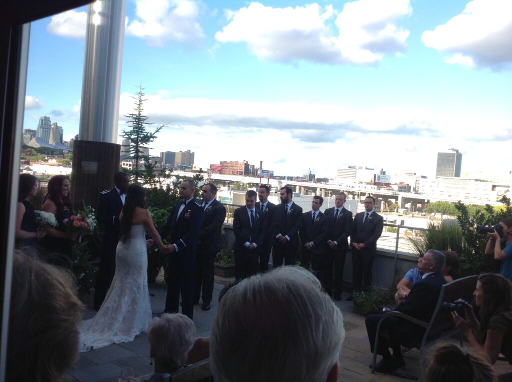 A bride and groom exchange vows outdoors surrounded by bridesmaids and groomsmen in formal attire, with a city skyline and blue sky with clouds in the background. Guests are seated and a photographer captures the moment.