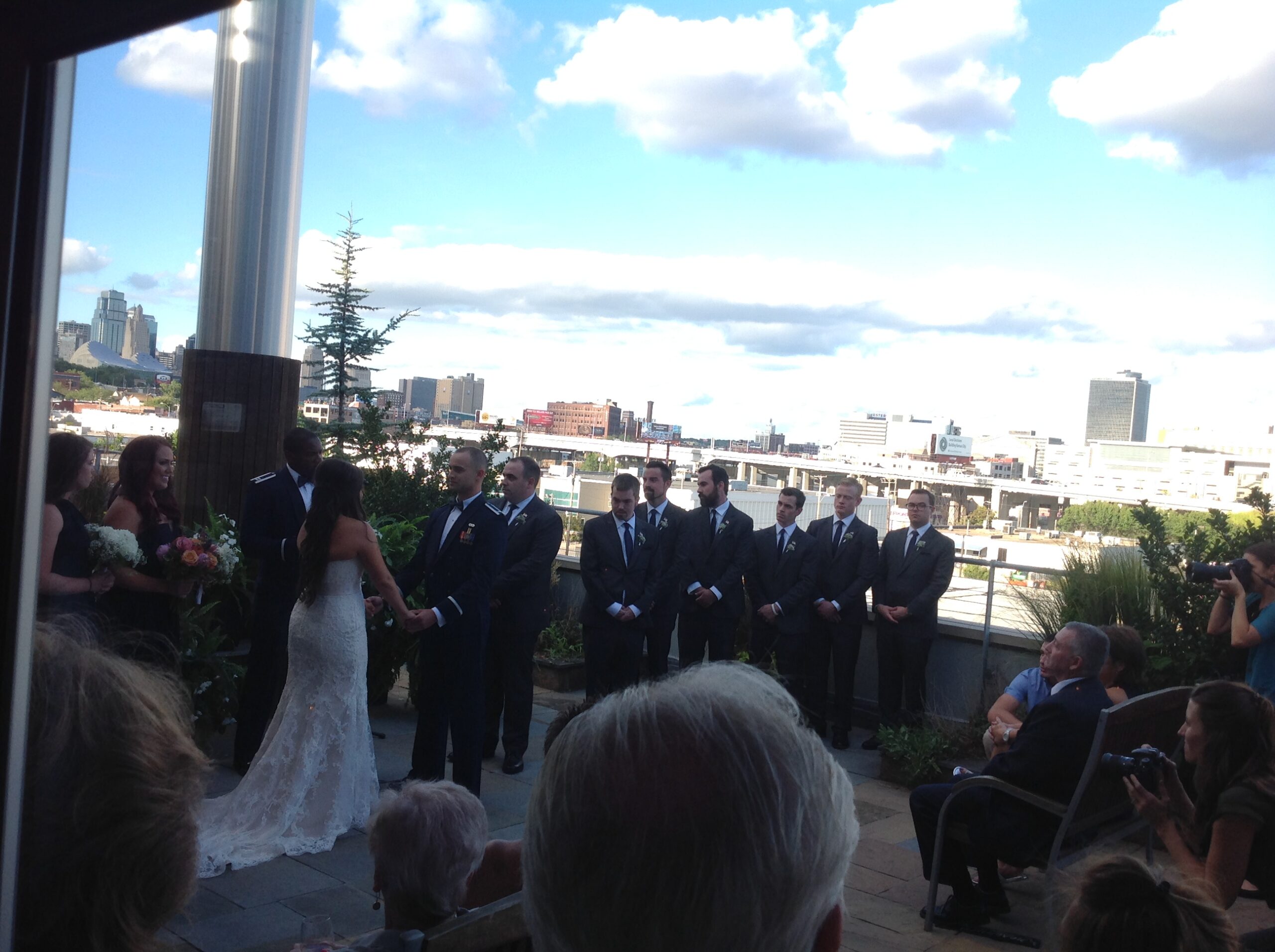 A bride and groom exchange vows outdoors surrounded by bridesmaids and groomsmen in formal attire, with a city skyline and blue sky with clouds in the background. Guests are seated and a photographer captures the moment.