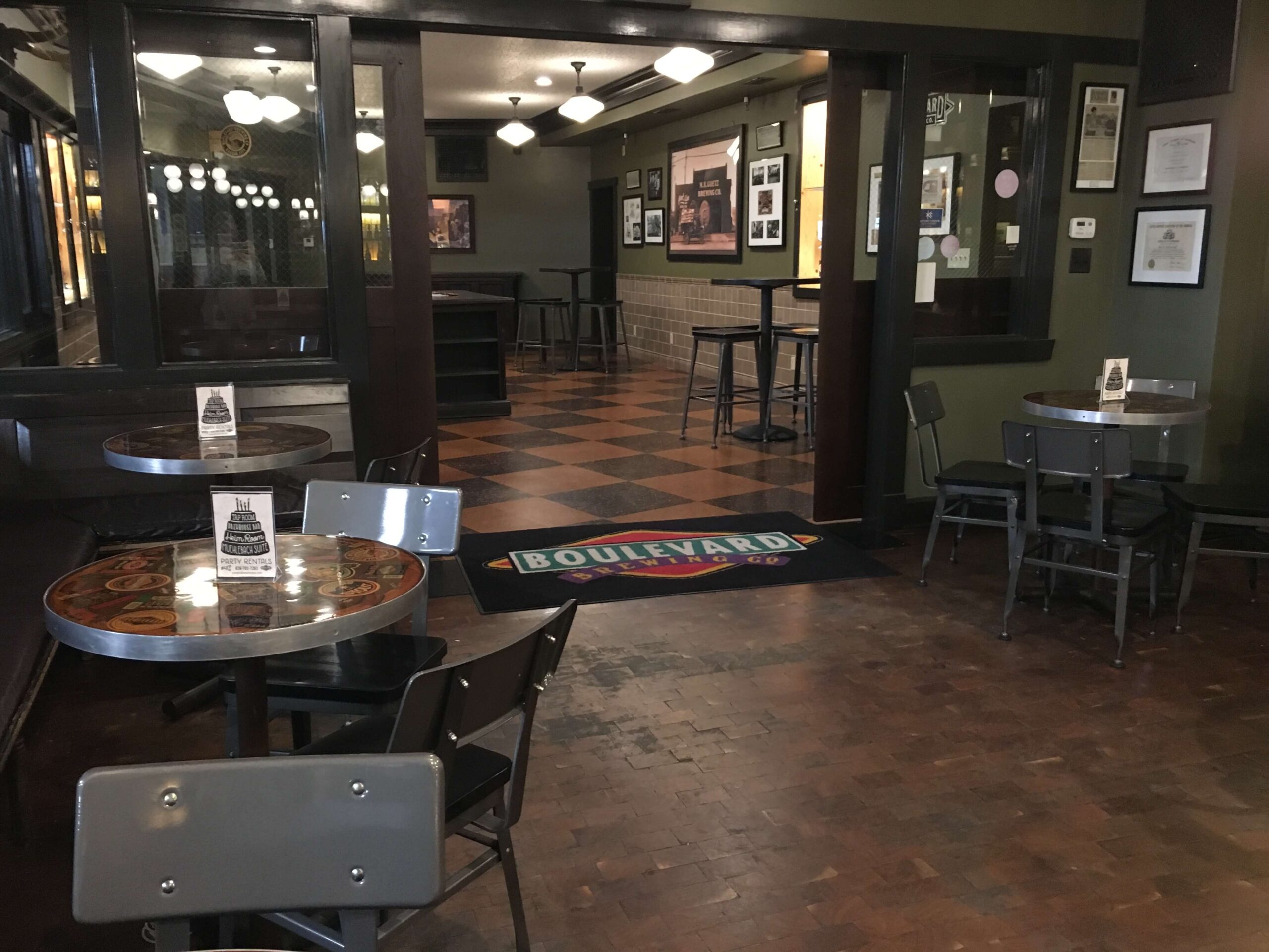 A cozy, empty bar with small round tables and metal chairs, dark wood floors, and framed pictures on green walls. A rug with the word "Boulevard" leads to a checkered floor area in the back.