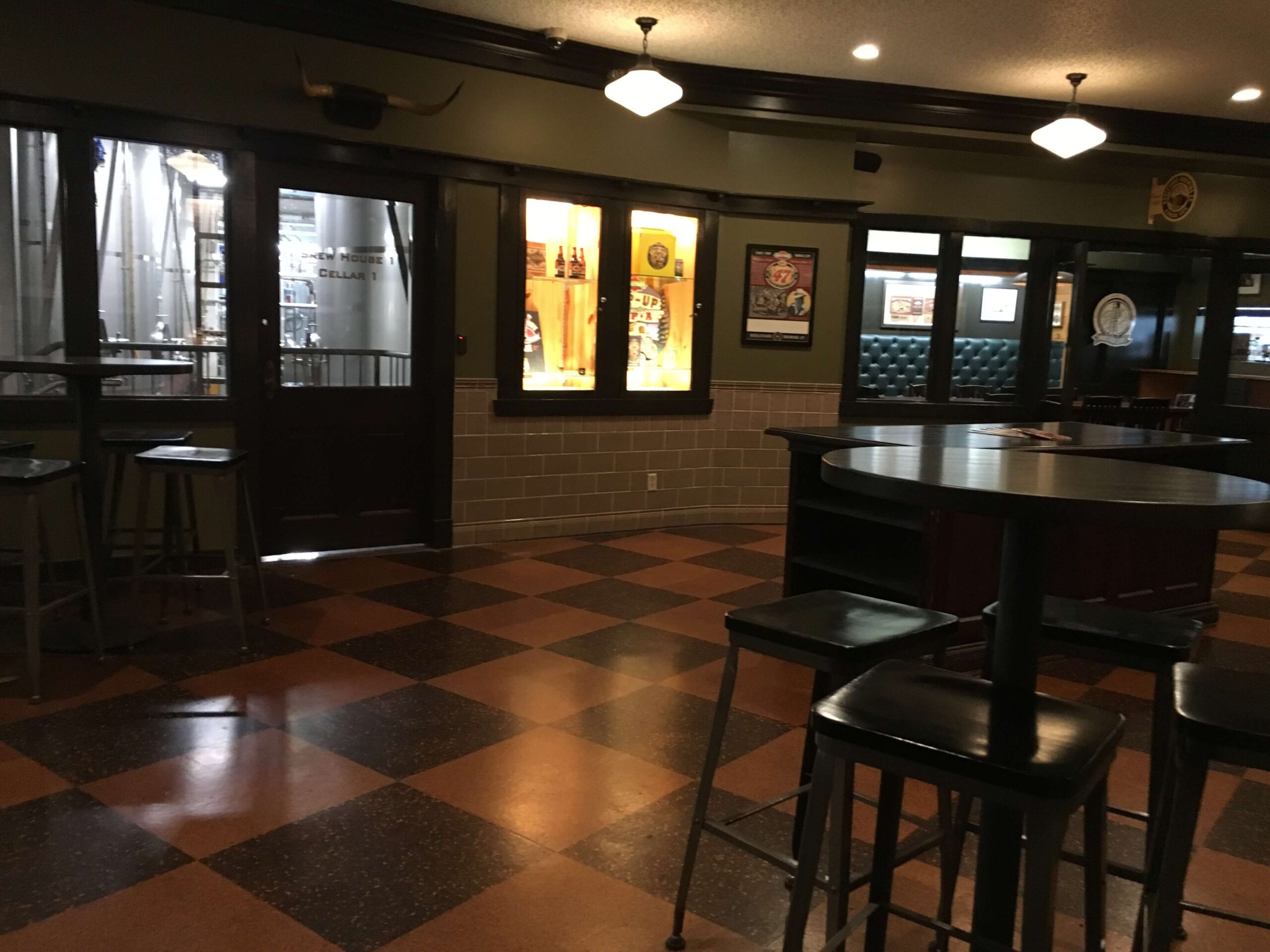 A dimly lit bar or pub interior with checkered tile flooring, high tables and stools, framed pictures on the wall, and windows revealing a brewery area in the background.
