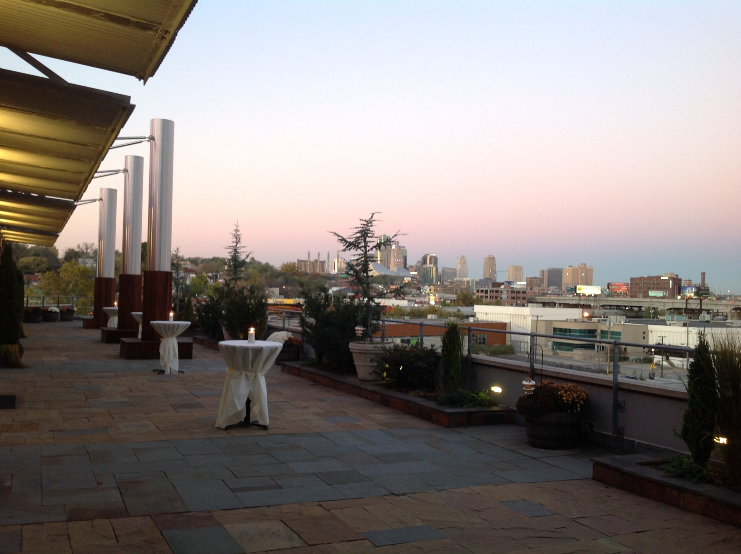A rooftop patio at sunset, with tall tables draped in white cloth, potted plants, and a view of a city skyline under a pastel sky. The patio is empty and lined with stone tiles.