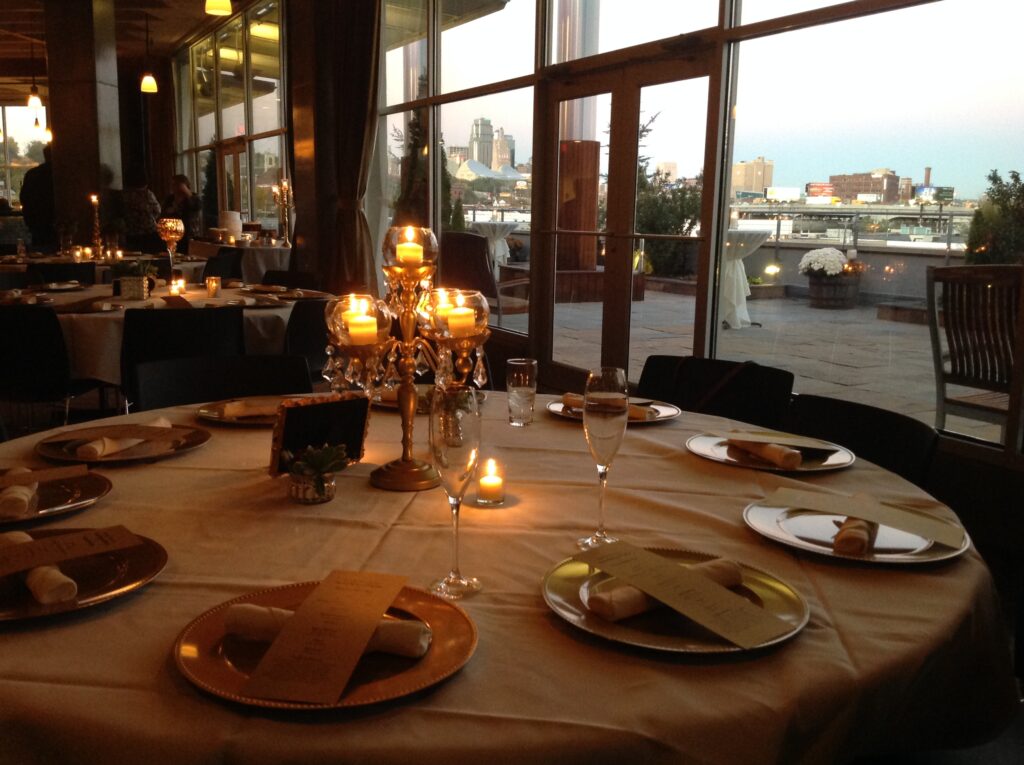 A round dinner table set with plates, napkins, menus, and a candelabra centerpiece with lit candles overlooks a cityscape through large windows at dusk.