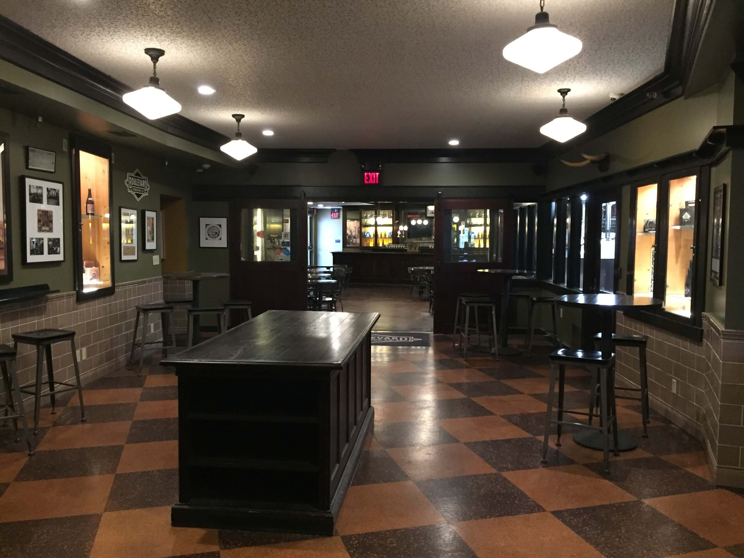 An empty bar or pub with dark wooden furniture, checkered brown tile floor, high tables and stools, framed pictures on the walls, and ceiling lights illuminating the space.