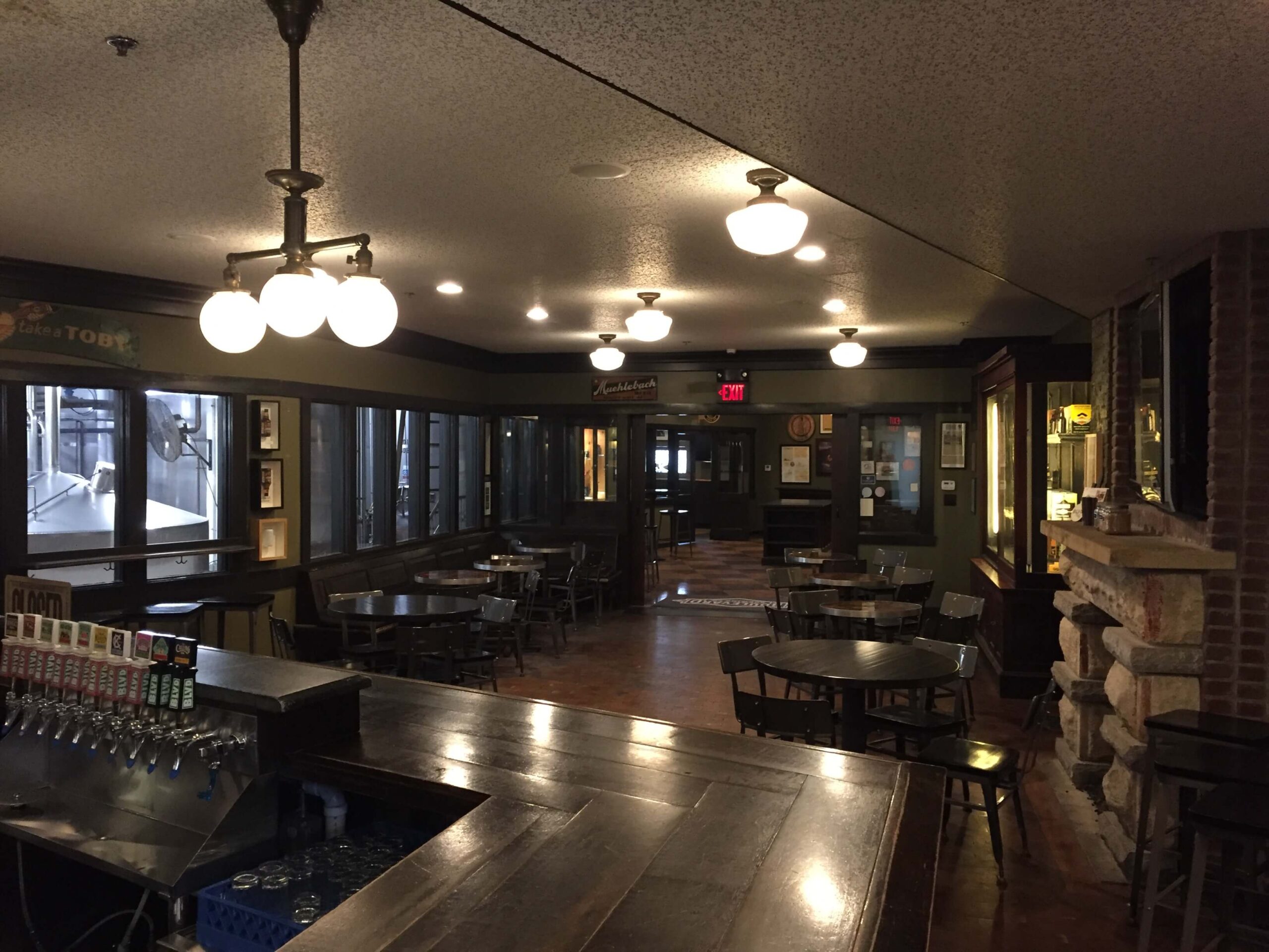 A dimly lit café or pub interior with round tables and chairs, hanging globe lights, a wooden bar with beer taps in the foreground, large windows, brick walls, and framed pictures on display.