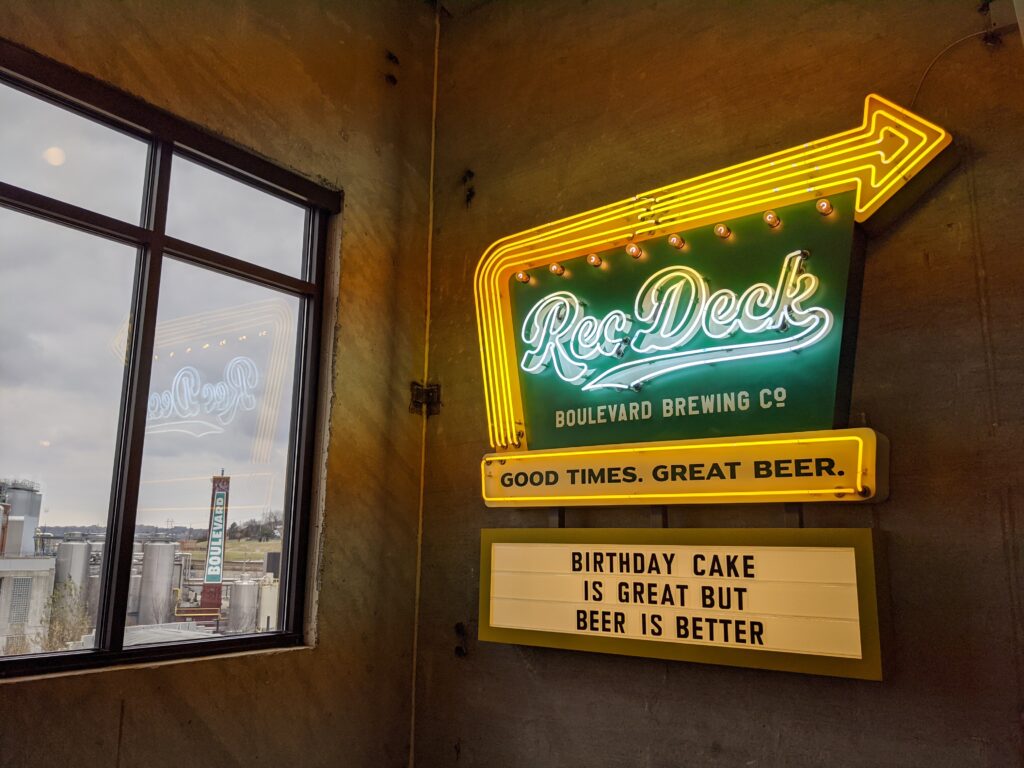 A neon sign on a wall reads “Rec Deck Boulevard Brewing Co. GOOD TIMES. GREAT BEER.” with a marquee below stating, “BIRTHDAY CAKE IS GREAT BUT BEER IS BETTER.” A window shows a city view outside.