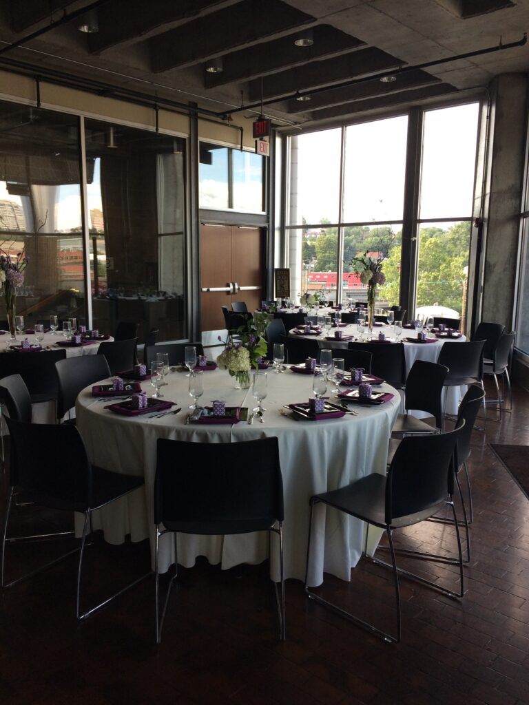 Round tables with white tablecloths, black chairs, and purple and white place settings are arranged in a modern room with large windows, letting in natural light and providing a view of trees outside.
