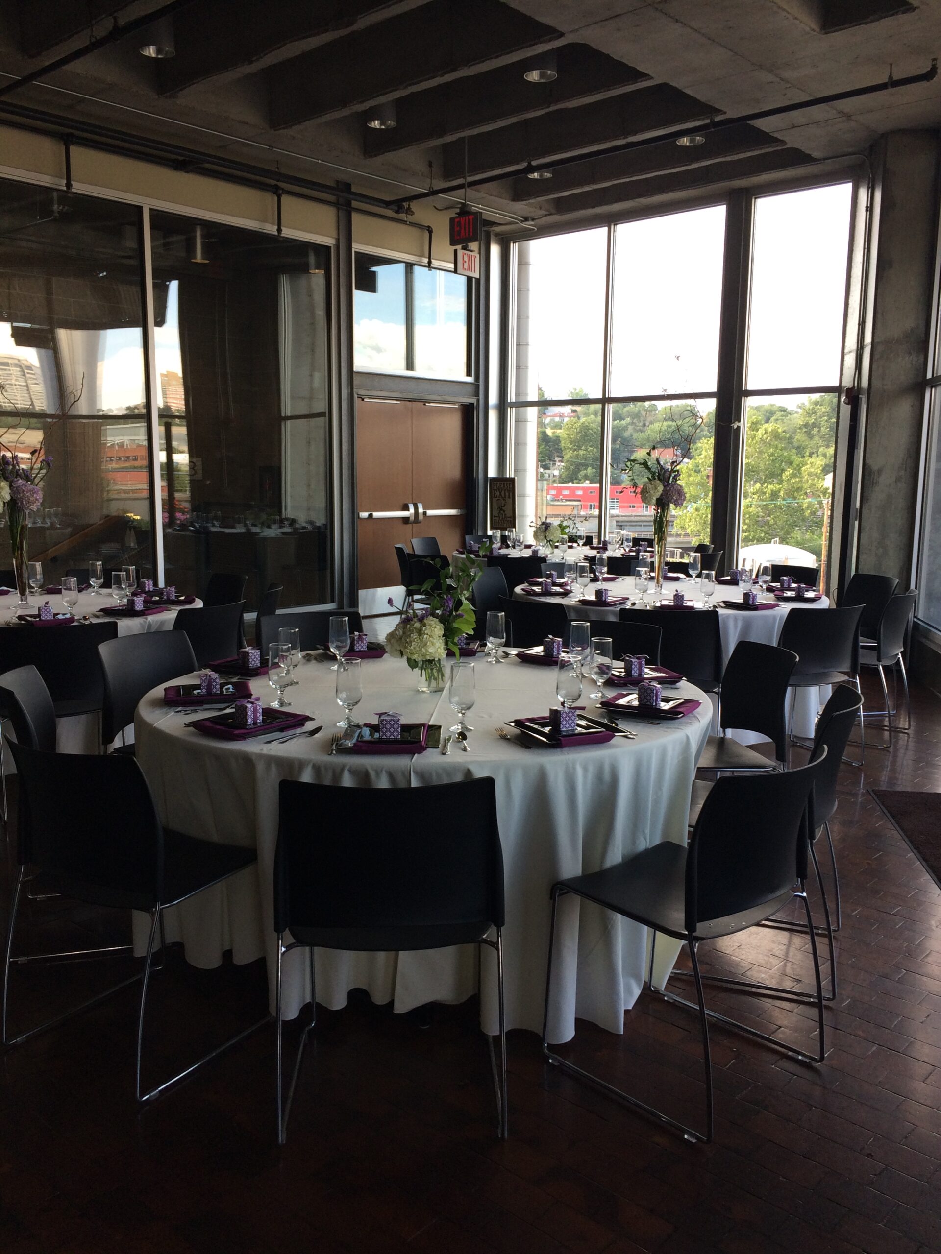Round tables with white tablecloths, black chairs, and purple and white place settings are arranged in a modern room with large windows, letting in natural light and providing a view of trees outside.