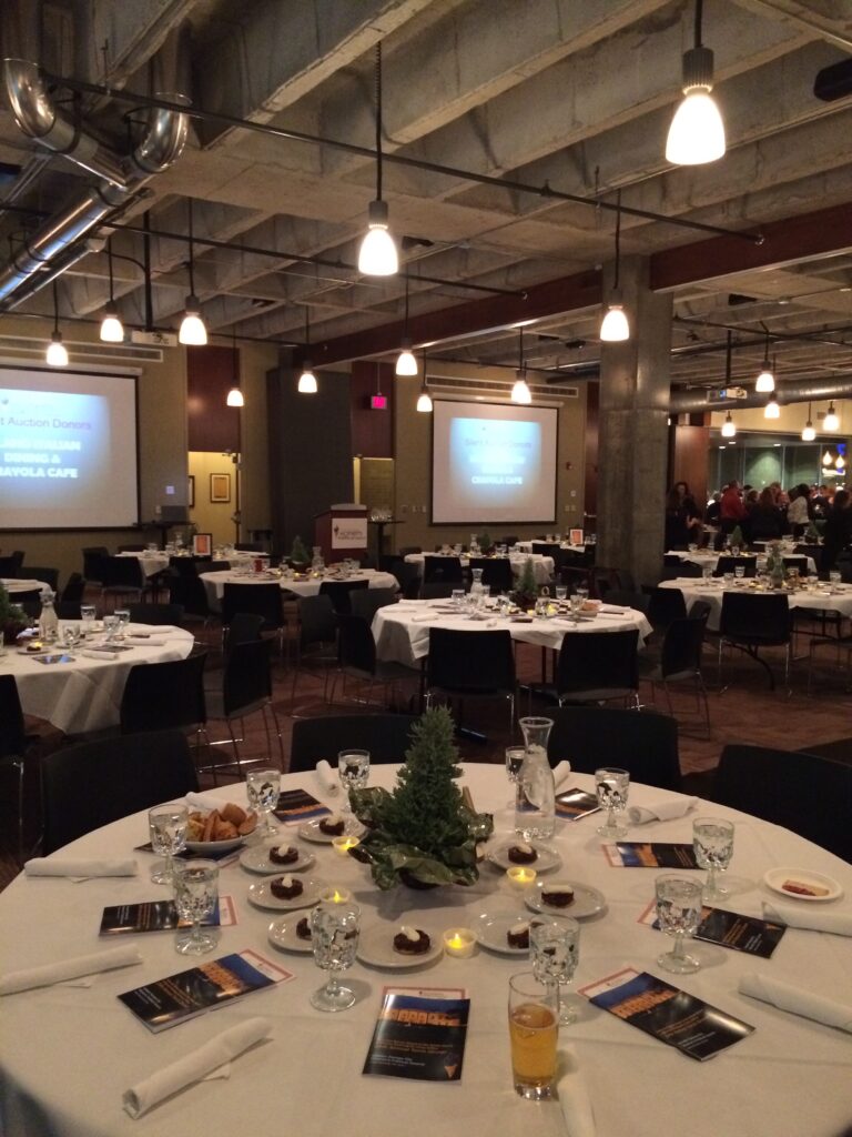 A banquet hall with round tables set for an event, featuring glasses, plates, programs, and small plants as centerpieces. Overhead lights illuminate the room, and presentation screens are visible in the background.