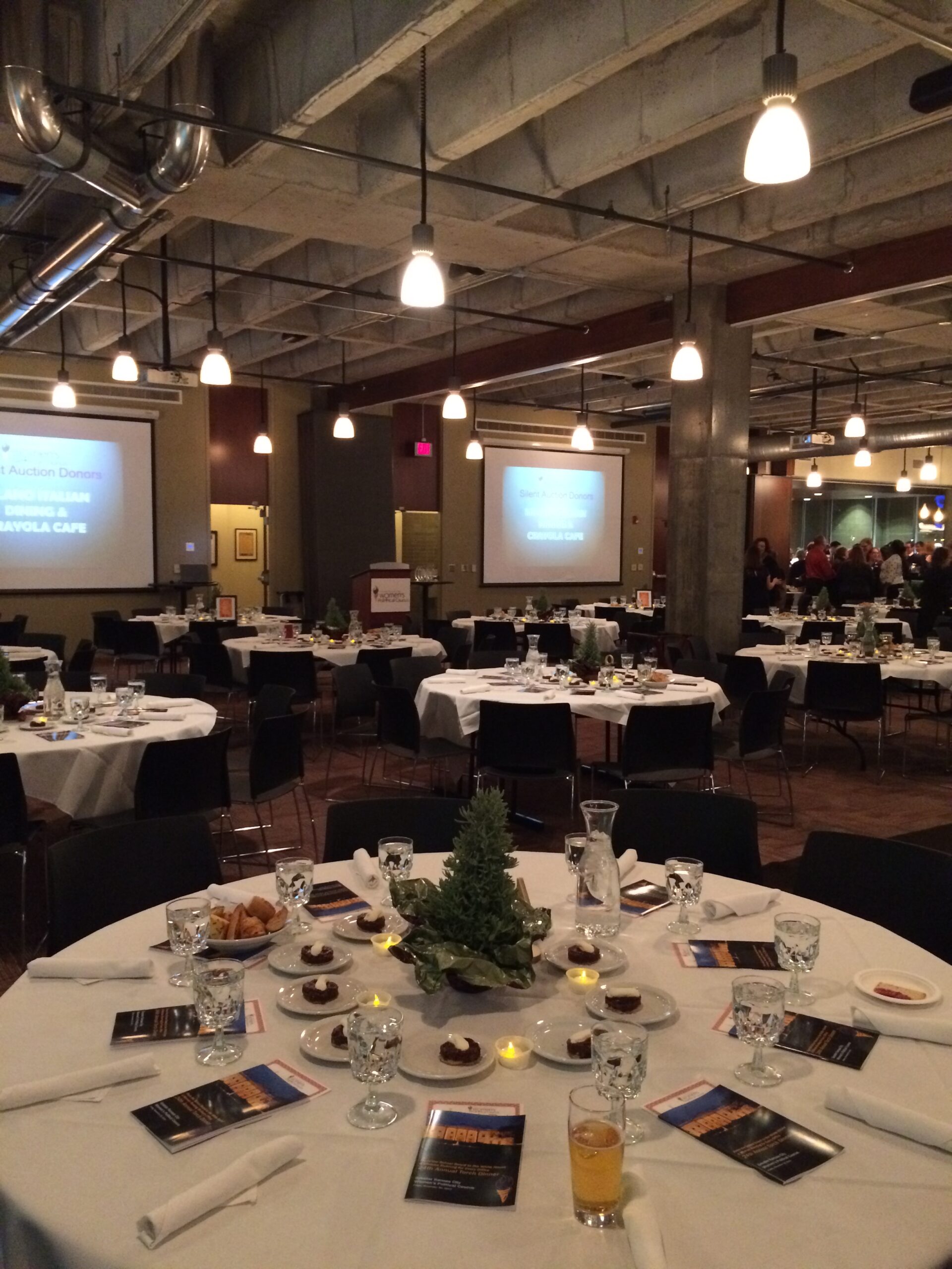 A banquet hall with round tables set for an event, featuring glasses, plates, programs, and small plants as centerpieces. Overhead lights illuminate the room, and presentation screens are visible in the background.