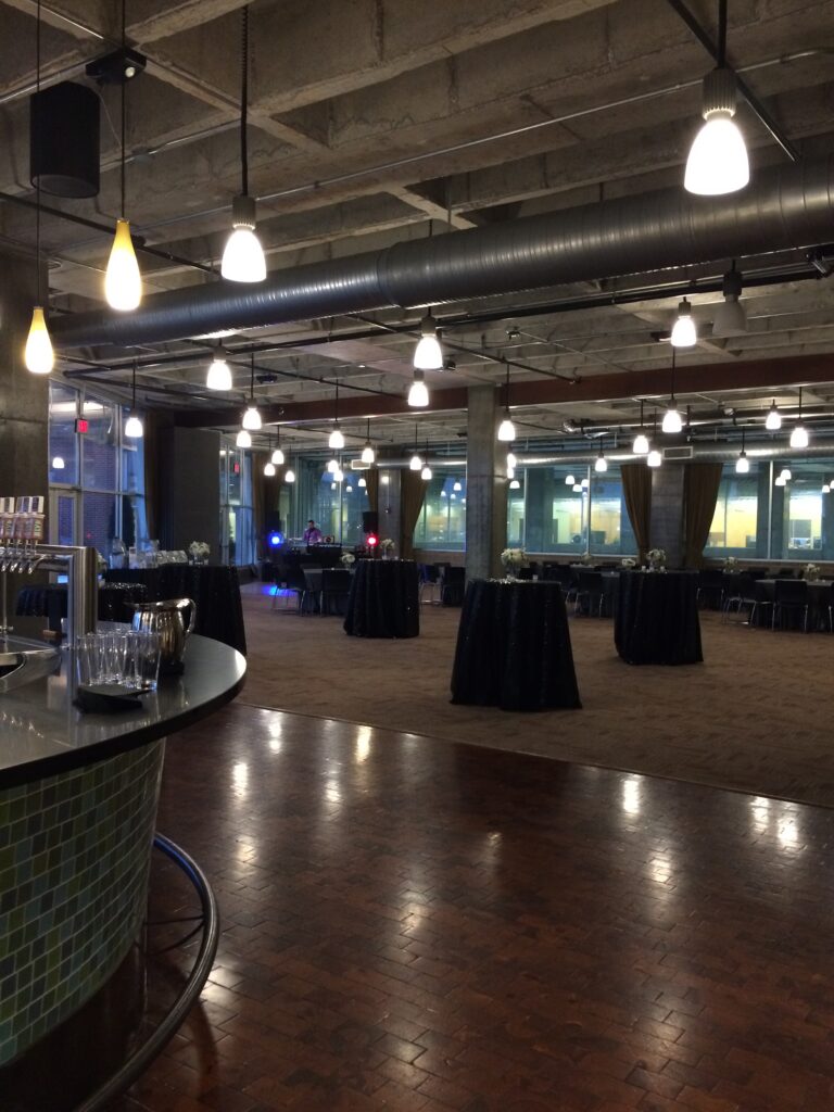 Spacious event hall with exposed concrete ceiling, hanging lights, round cocktail tables covered in black cloth, and large windows. Glasses and drinks are set up on a counter in the foreground.