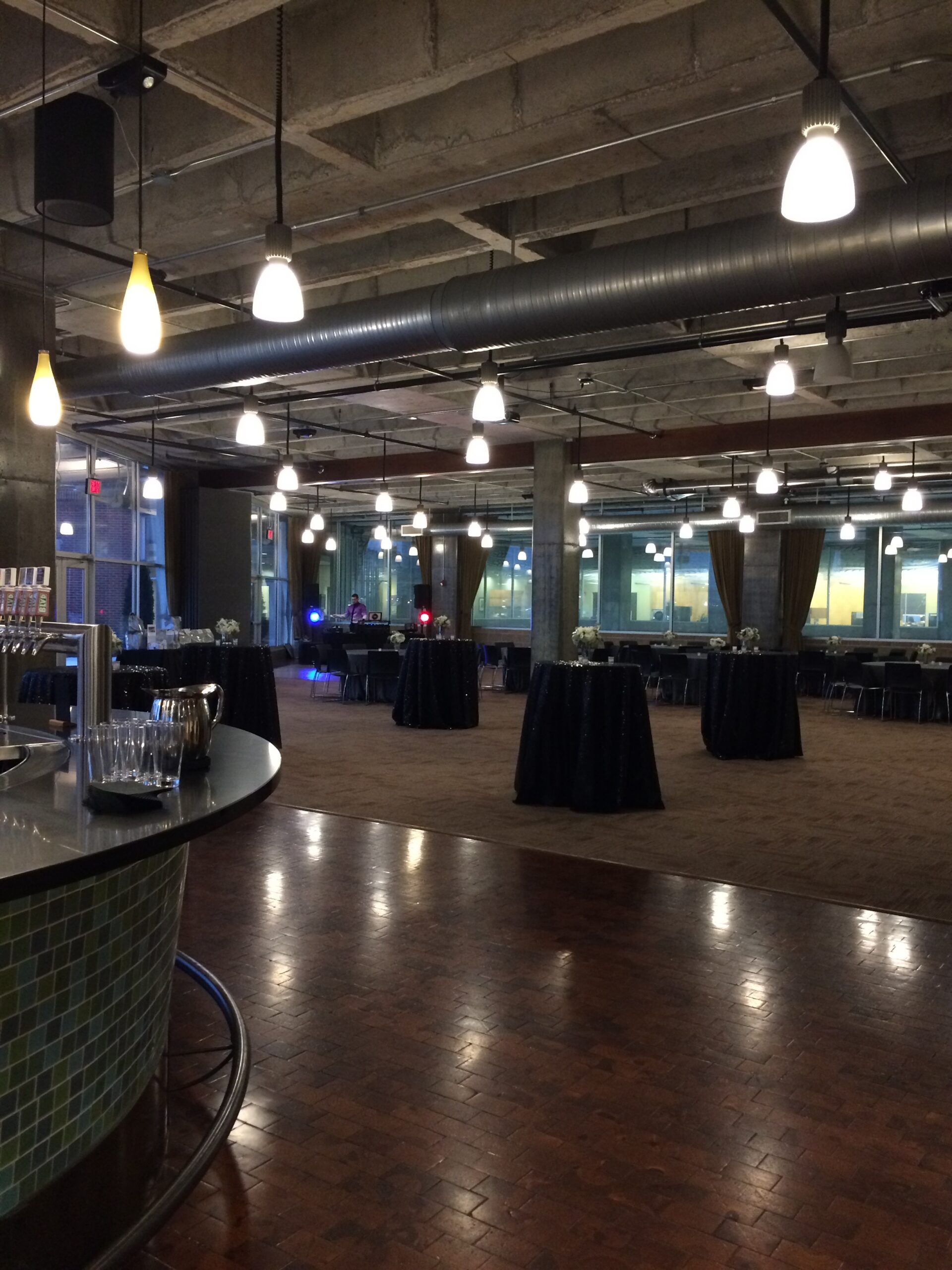 Spacious event hall with exposed concrete ceiling, hanging lights, round cocktail tables covered in black cloth, and large windows. Glasses and drinks are set up on a counter in the foreground.