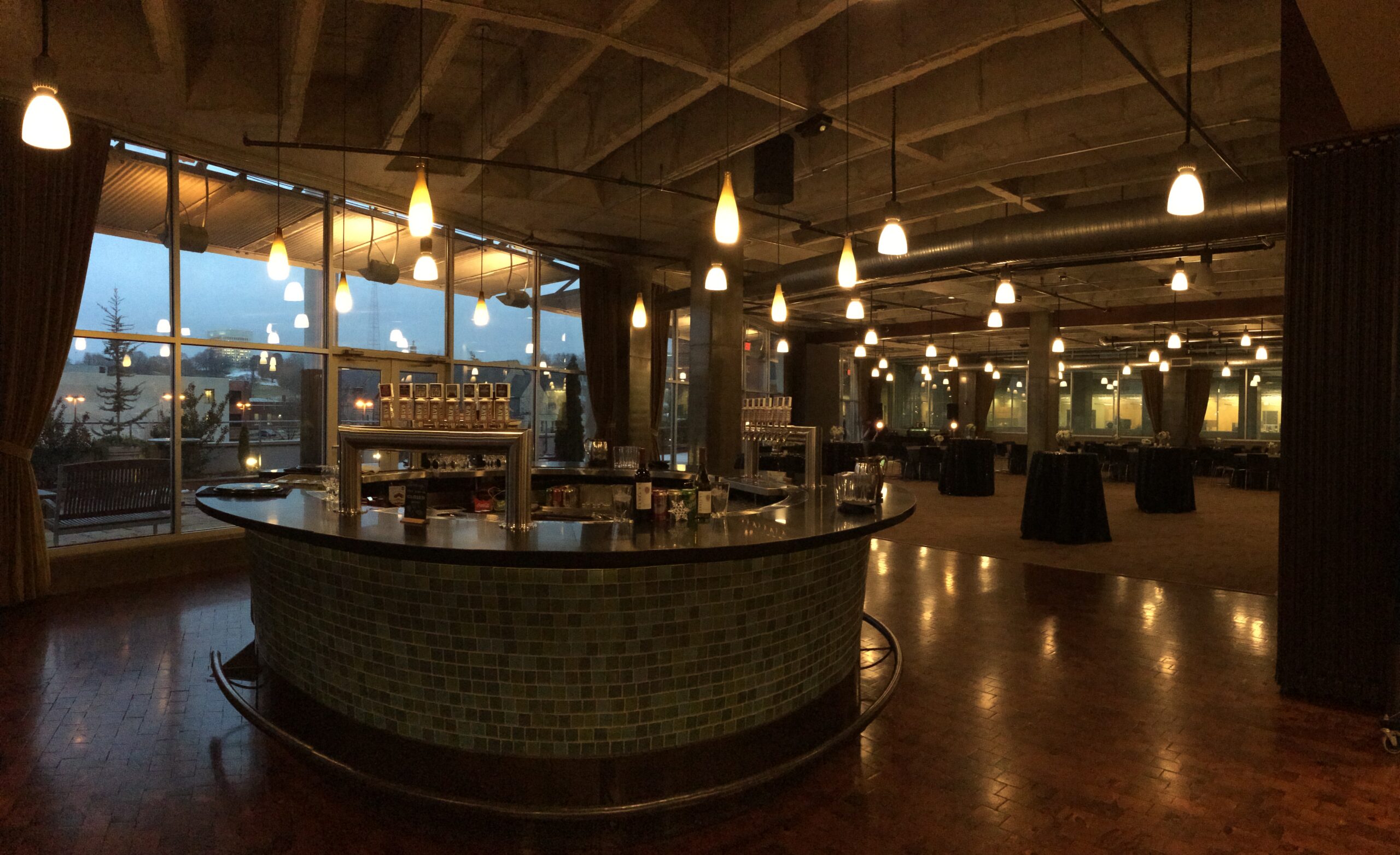 A dimly lit, modern event space with a central circular bar, hanging pendant lights, large windows, and empty round tables, set for an event during dusk.