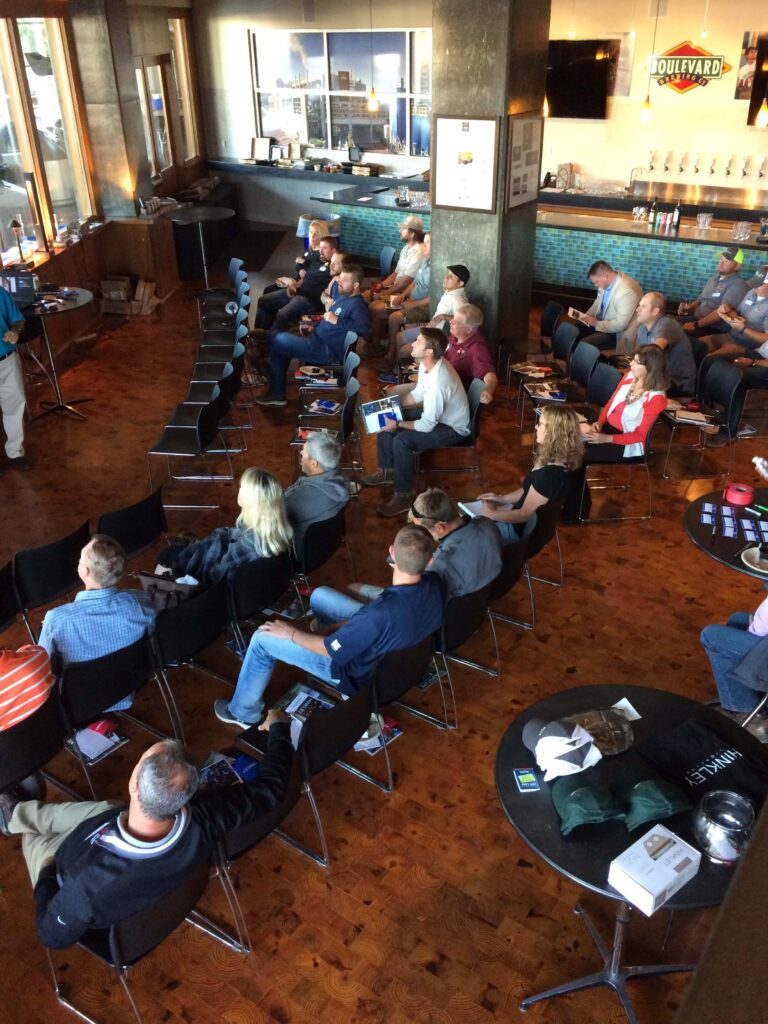 A group of people sit in rows of chairs facing a speaker in a modern room with large windows, a bar area in the back, and tables with merchandise or promotional items.