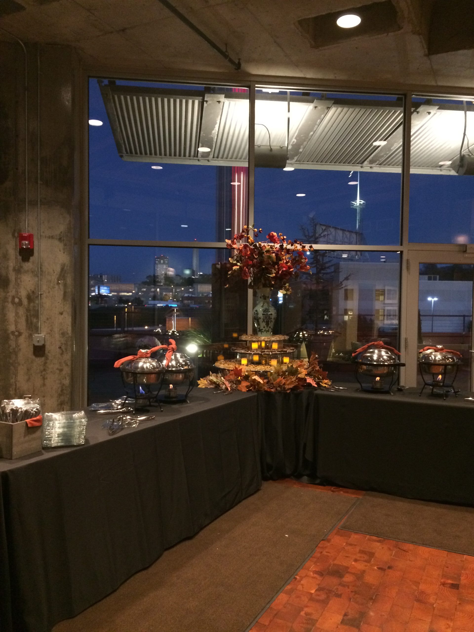 A buffet table with chafing dishes and autumn-themed decorations stands near large windows overlooking a cityscape at dusk, with lights visible outside.