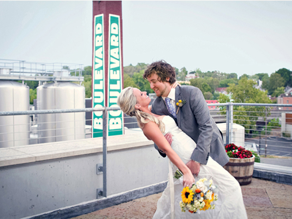 A groom in a gray suit dips his smiling bride in a white dress as they pose on a rooftop, holding a bouquet of sunflowers, with a "Boulevard" sign and tanks in the background.