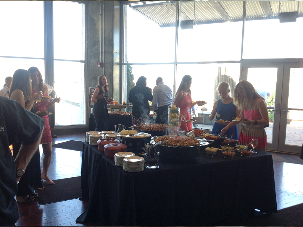 A group of people serving themselves food from a buffet table in a well-lit room. The table is filled with various dishes and plates. Large windows are in the background, allowing natural light to fill the space.