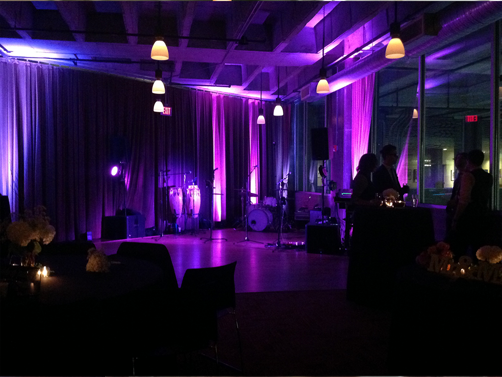 Dimly lit room with purple lighting, featuring a small stage with musical instruments including drums and congas. Several people stand near cocktail tables, and pendant lights hang from the ceiling.