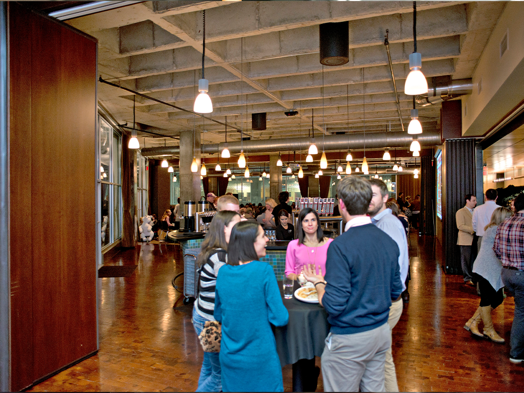 A group of people gather around a table in a large room with industrial ceilings and wooden floors. Overhead lights illuminate the space. More people are visible in the background, engaged in conversation.