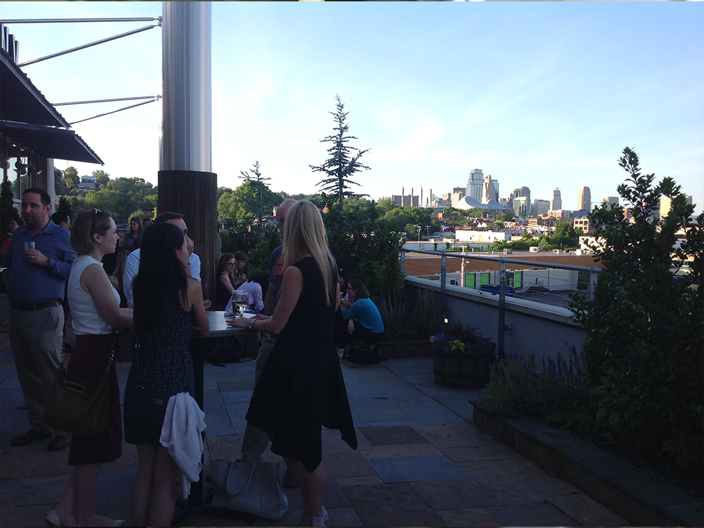 A group of people socializing on a rooftop patio during the day. The city skyline is visible in the background. Some are standing around tables, and trees are scattered around the area. The scene is casual and lively.