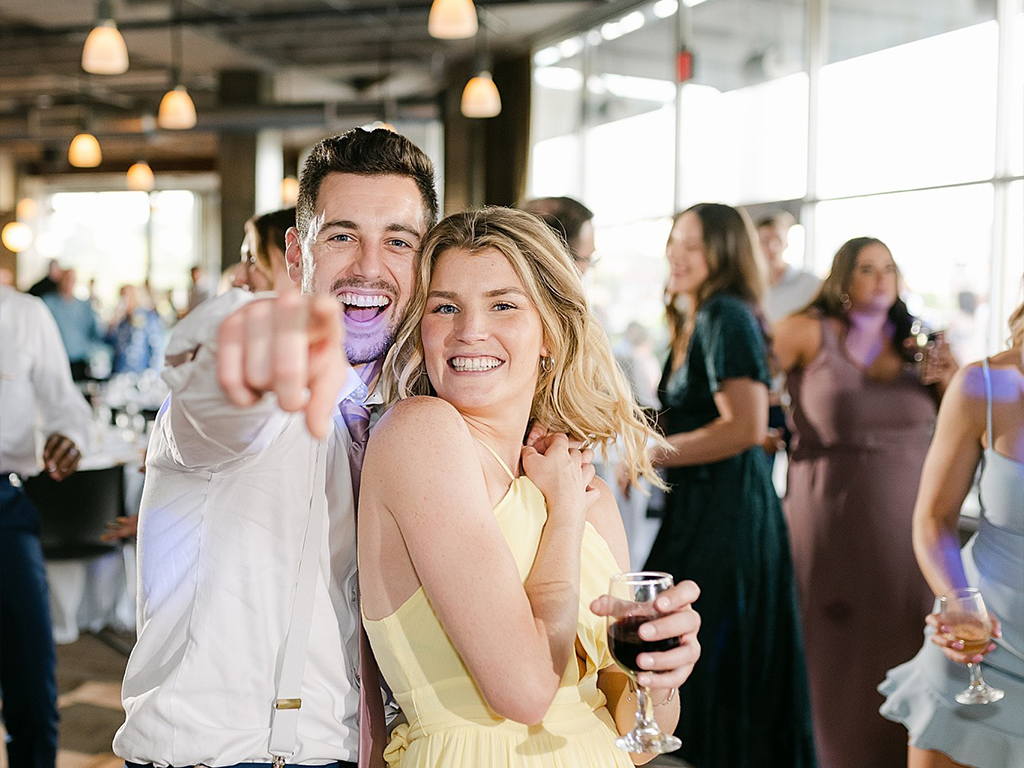A smiling man and woman, with the man pointing towards the camera. The woman in a yellow dress holds a glass of red wine. They appear to be at a lively event or celebration with other people in the background.