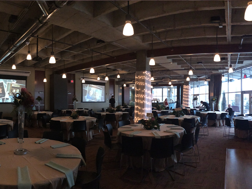 A dimly lit event space with round tables covered in white tablecloths and arranged with chairs. The room is decorated with string lights wrapped around columns. A projector displays images on the wall. High ceilings show exposed pipes.