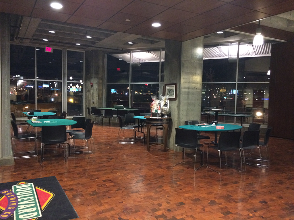 A room with several round tables covered in green cloth, surrounded by black chairs. Large windows reveal a cityscape at night. A floor mat with "Boulevard" lies in the entrance. A gift basket is placed on a table.