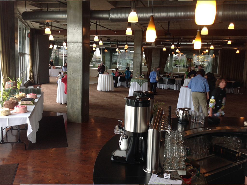 A spacious event hall with tables covered in white tablecloths, each displaying assorted cakes. A few people are mingling in the background. The room has industrial-style lighting and large windows. A coffee station is set up in the foreground.