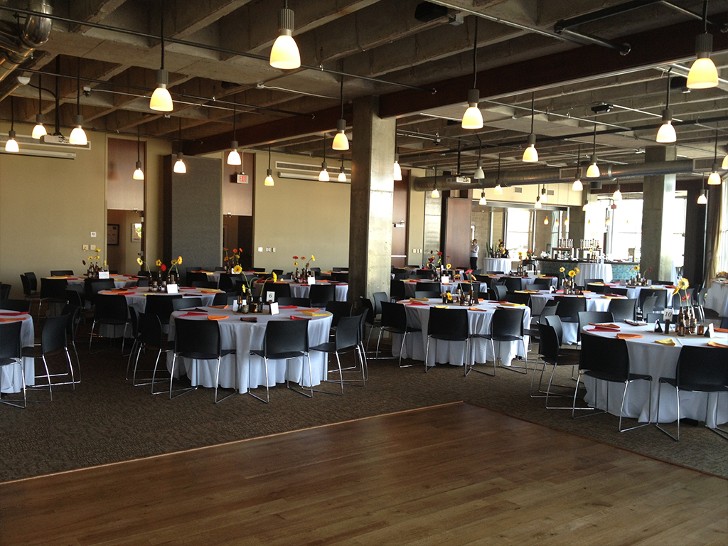 A spacious event hall with round tables covered in white tablecloths, each adorned with floral centerpieces. Black chairs surround the tables. Warm lighting from hanging lamps creates an inviting atmosphere.