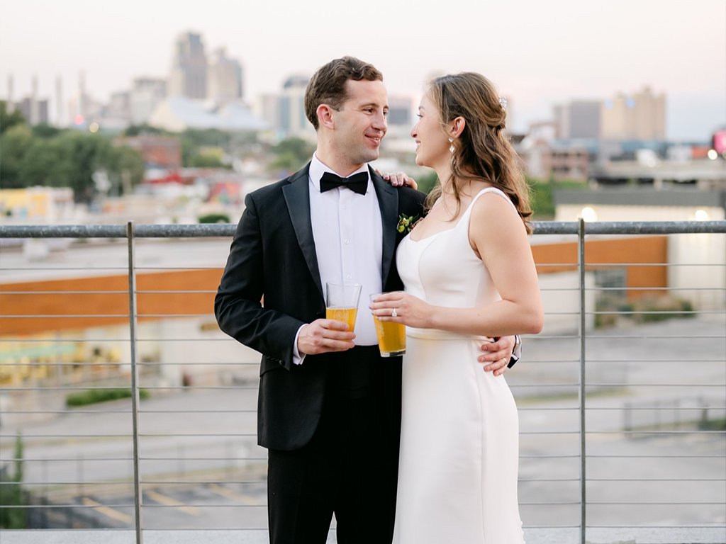 A couple in formal attire stands on a rooftop, holding drinks and gazing at each other. The man is in a black tuxedo and the woman in a white dress. The city skyline and buildings are visible in the background.