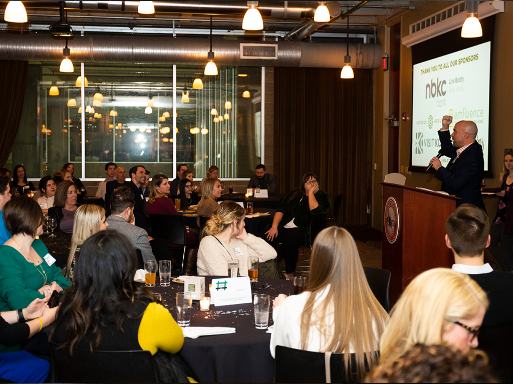 A speaker addresses a seated audience in a dimly lit event space. Tables are set with drinks and paper programs. The screen displays a presentation with logos. Ceiling lights and exposed pipes add to the room's industrial feel.