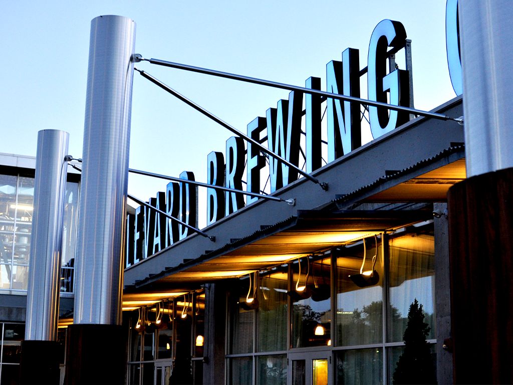 View of a brewing company entrance featuring large metallic columns and a sign with bold letters spelling "BREWING." The entrance is illuminated with warm lights, and the sky is clear.