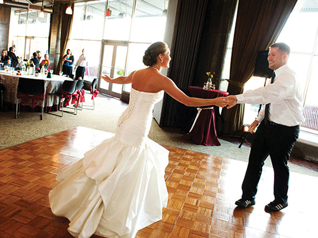 A bride in a white gown and a groom in a shirt and tie are dancing on a wooden floor. Guests are seated at round tables in the background, and large windows let in natural light. The bride is spinning, holding the groom's hand.