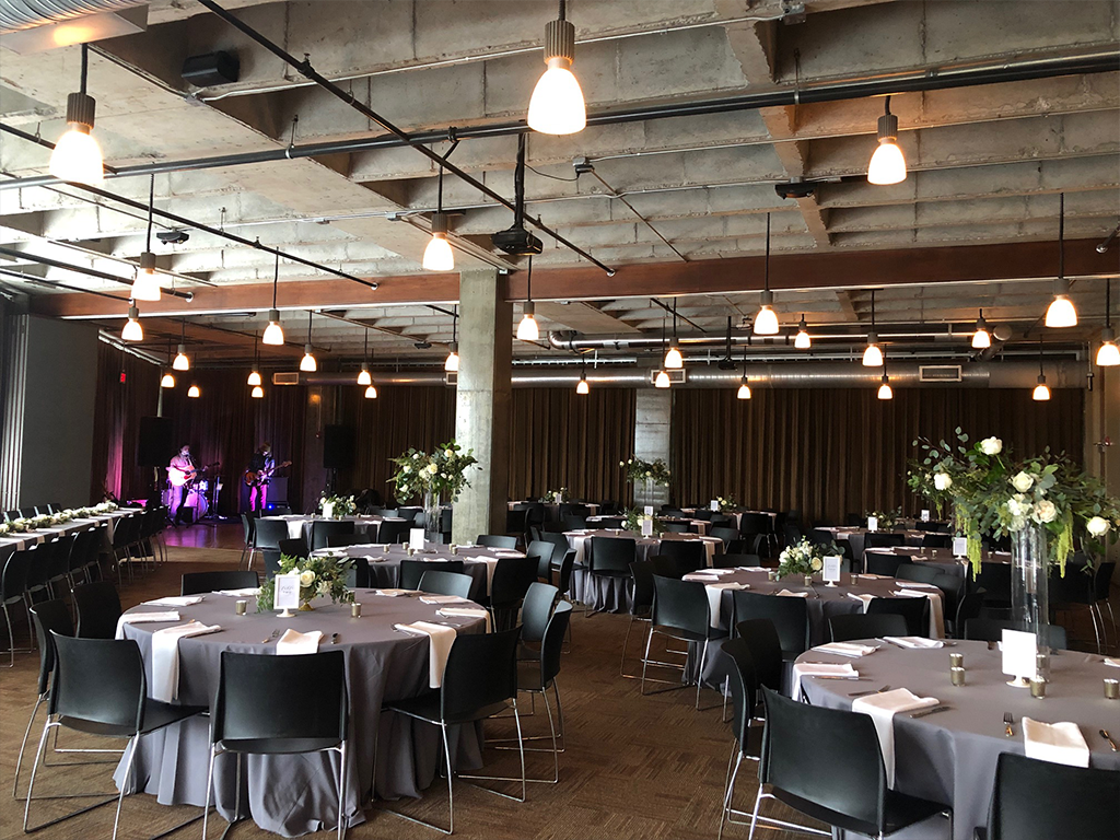 Spacious event venue with round tables covered in gray tablecloths and black chairs. Each table has floral centerpieces. Stage with musicians in the background. Ceiling features industrial lighting and exposed ducts, creating a modern ambiance.