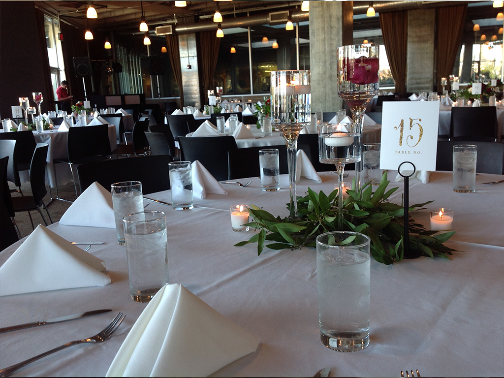A dining setting with tables covered in white cloths, folded napkins, glasses of water, and small candles. Each table has a centerpiece of greenery and a table number sign. Warm lighting hangs from the ceiling, creating an elegant atmosphere.