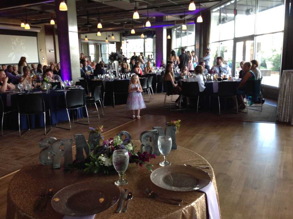 A young girl in a purple dress stands alone in front of a decorated sweetheart table at a wedding reception, with guests seated at round tables in a modern, spacious venue.