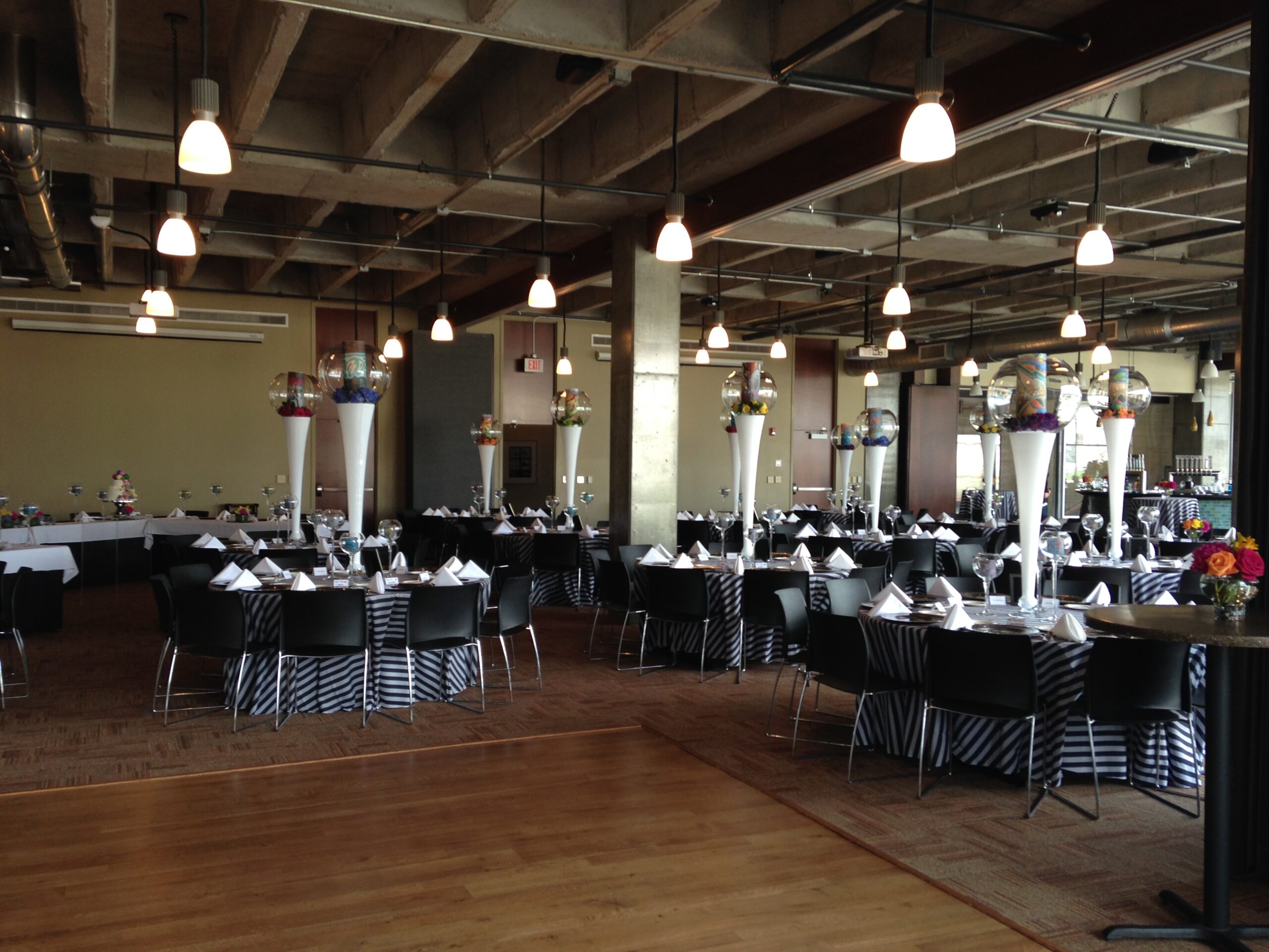 A modern event space with round tables covered in black-and-white striped linens, each set with glassware and white napkins. Tall centerpieces with balloons decorate the tables, and pendant lights hang from the ceiling.