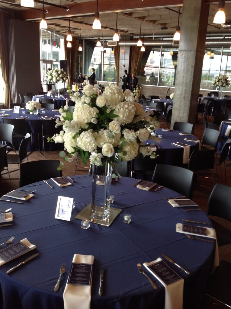 Round tables with navy blue tablecloths are set for an event, each featuring a tall glass vase with white flower arrangements, menu cards, and neatly arranged silverware in a modern, well-lit venue.