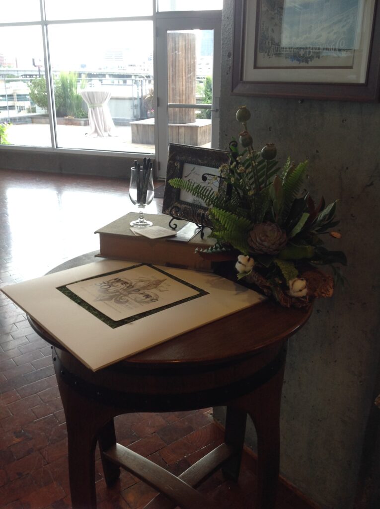 A round wooden table by a window holds a framed photo, a guestbook, stacked books, a pen in a holder, and a floral arrangement. City buildings and a patio are visible through large windows in the background.