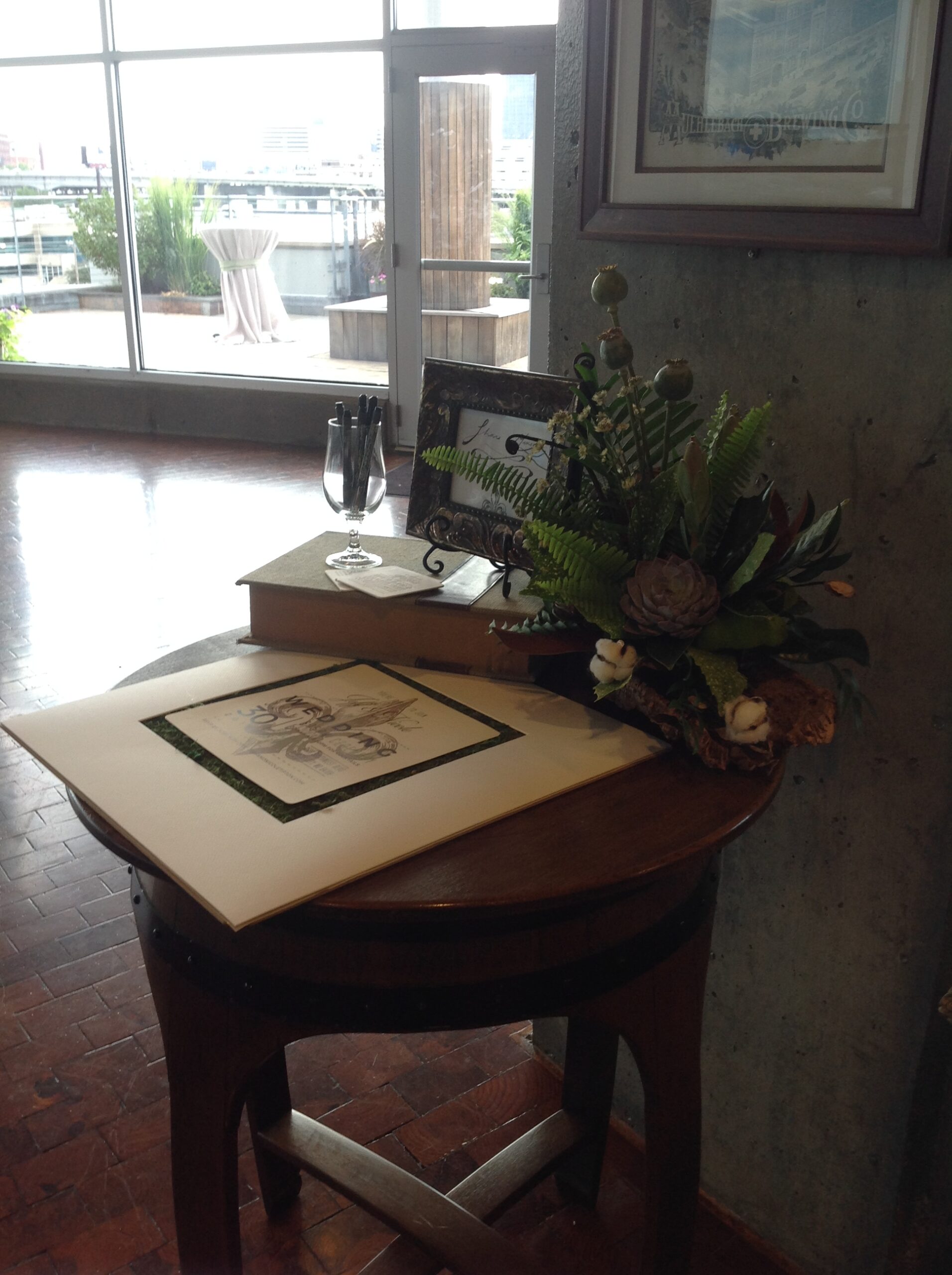 A round wooden table by a window holds a framed photo, a guestbook, stacked books, a pen in a holder, and a floral arrangement. City buildings and a patio are visible through large windows in the background.