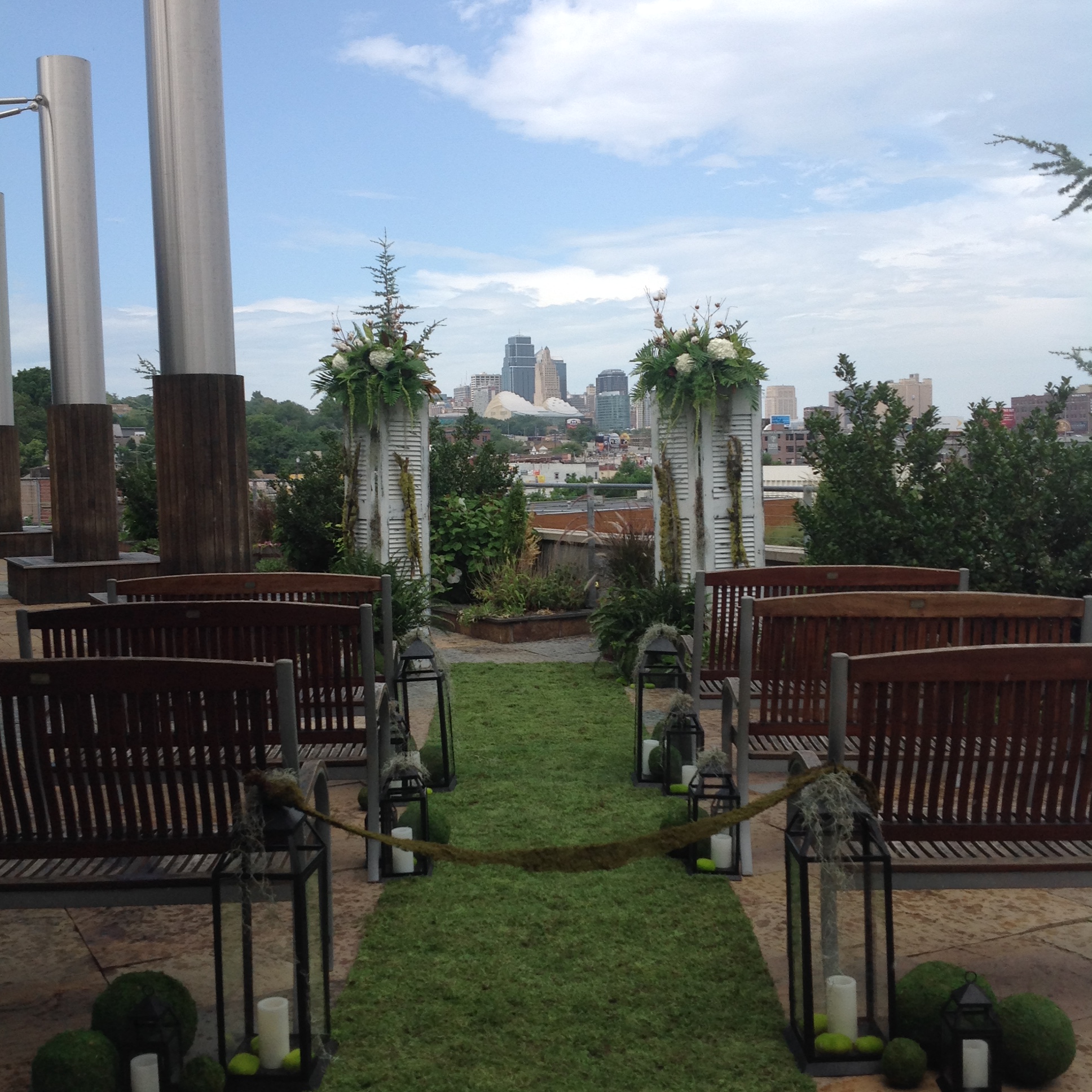 Outdoor wedding ceremony setup with wooden benches facing a green aisle lined with lanterns and floral arrangements, overlooking a city skyline under a partly cloudy sky.